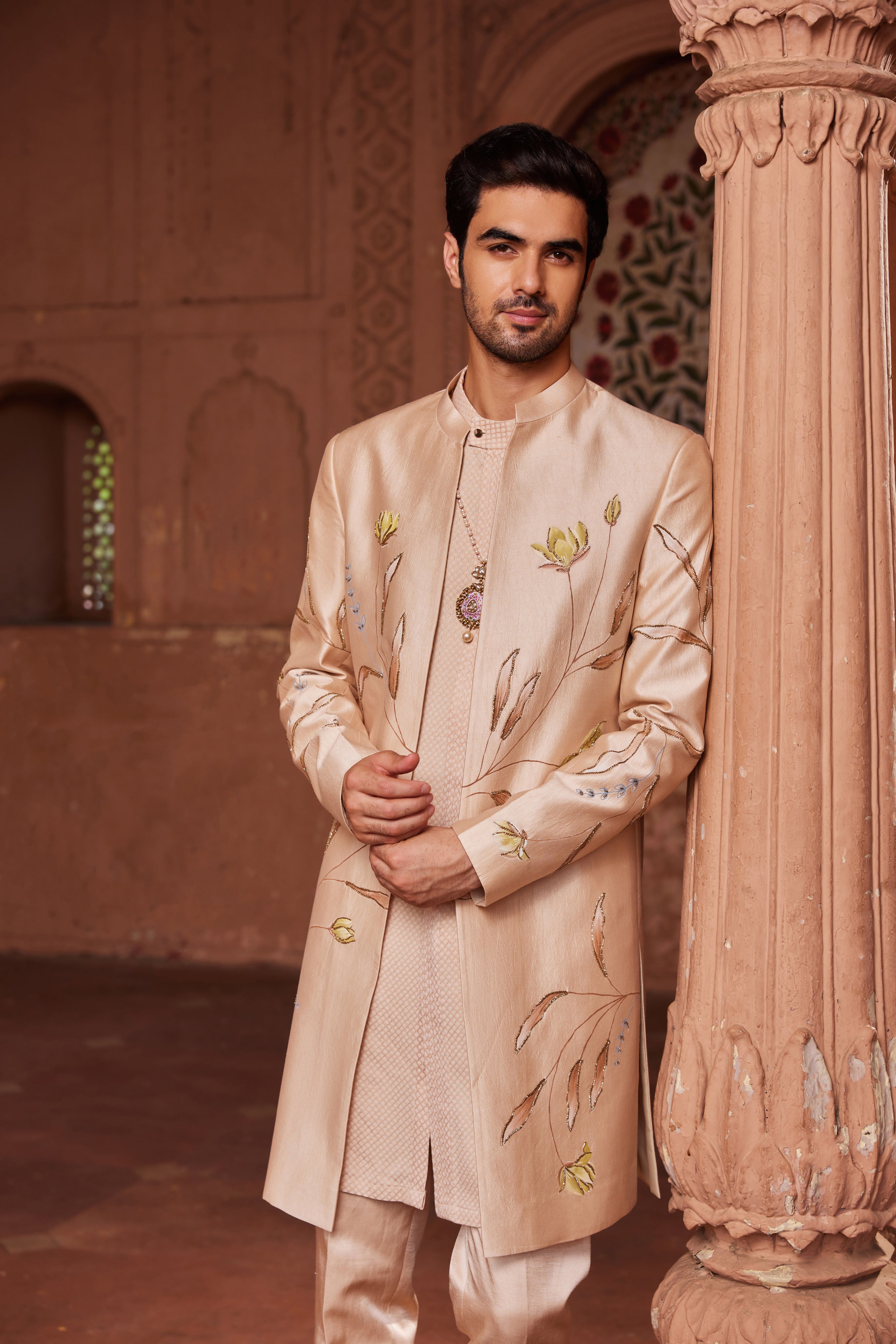 The Bageecha Open Sherwani Set showcases a sherwani adorned with meticulously hand-painted designs and detailed dabka and zardozi embellishments. It is complemented by a simple, monotone kurta and pants