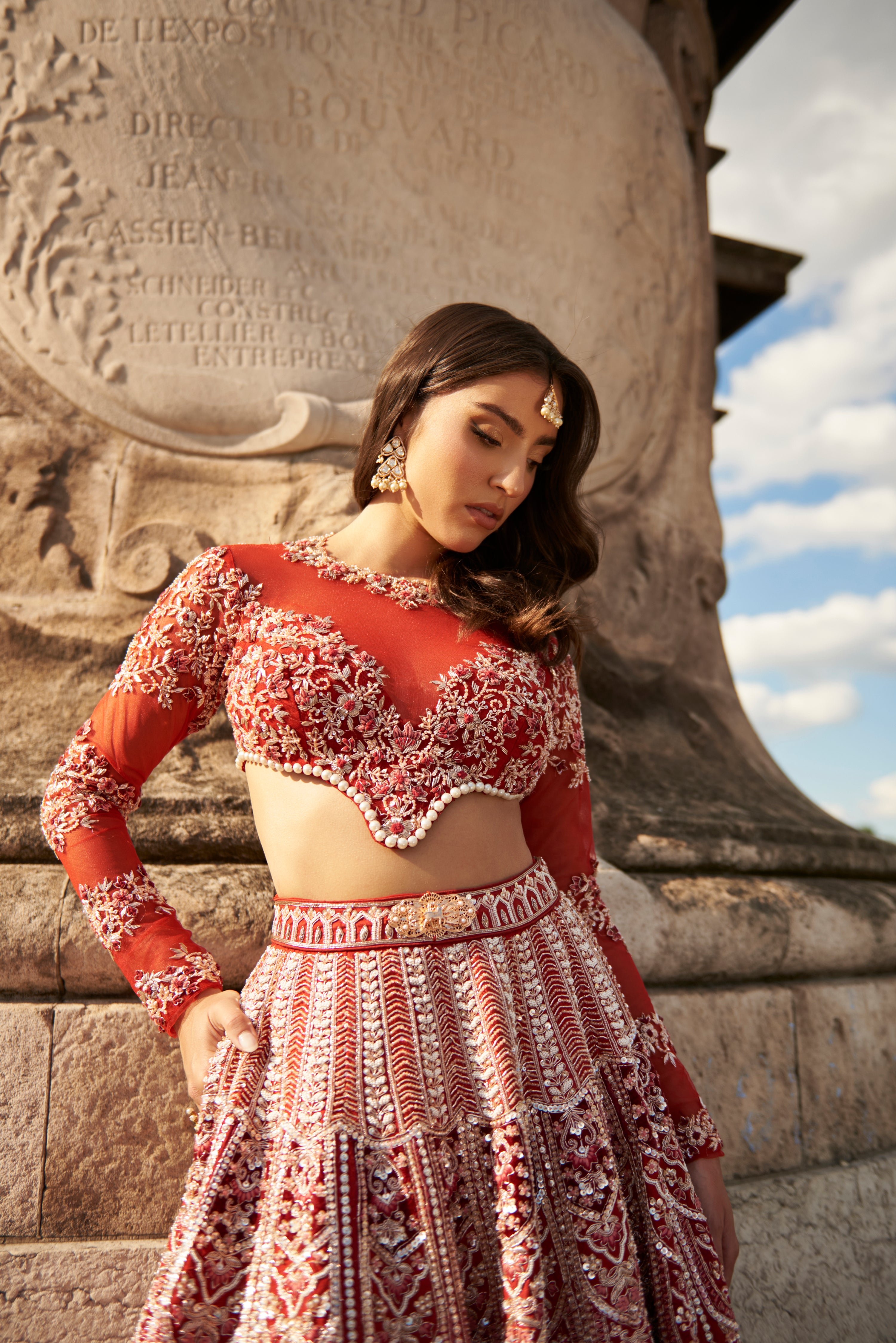 Full-sleeve red lehenga set in carnelian red, featuring intricate hand embroidery and pearl embellishments, crafted from luxurious raw silk and velvet, paired with a sheer net full-sleeve blouse and dupatta