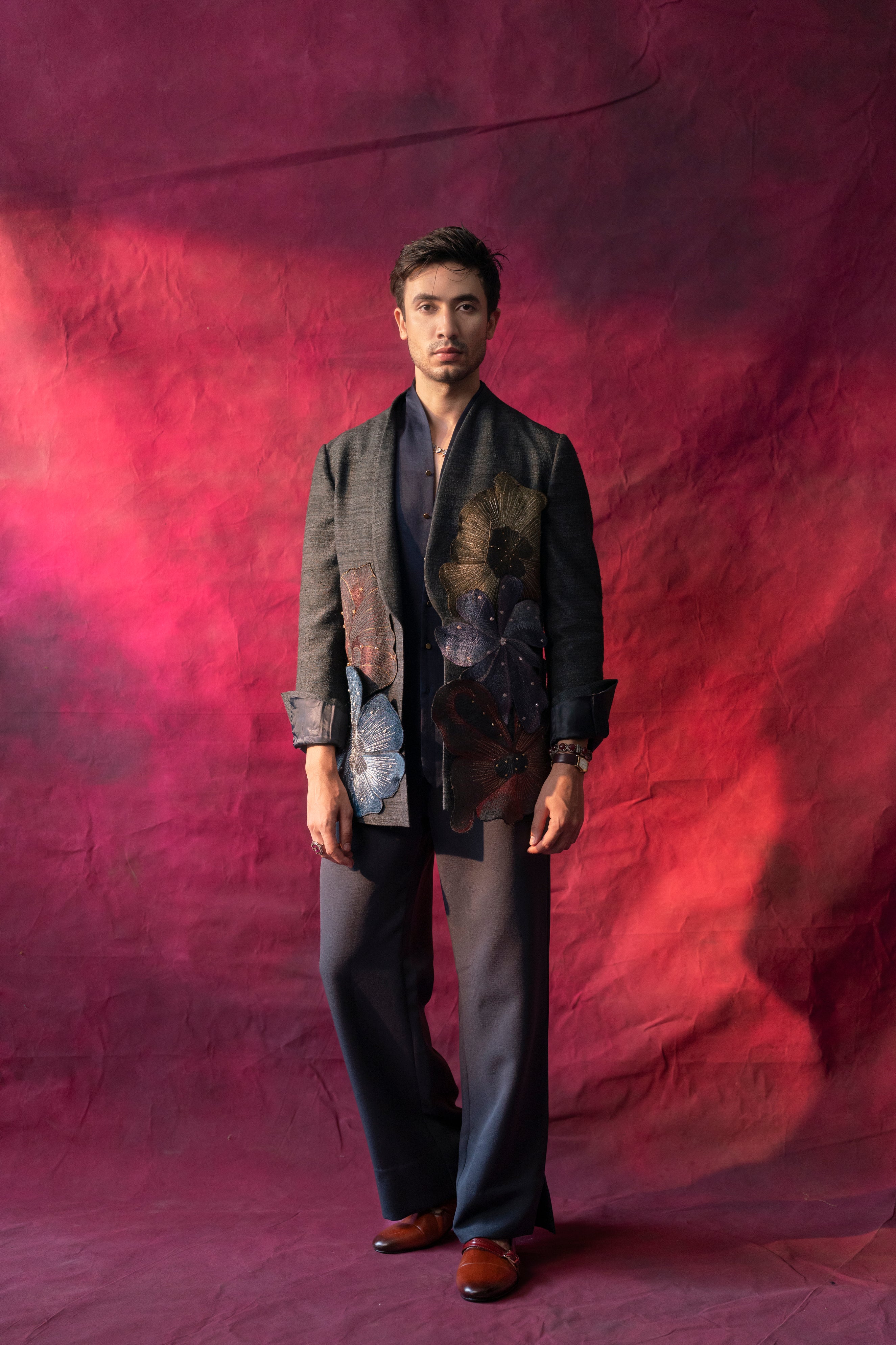 A stylish ensemble featuring a raw silk blazer in charcoal grey, adorned with multi-color hand-embroidered flowers placed in an abstract design. Paired with a linen silk shirt and matching trousers for a refined look.