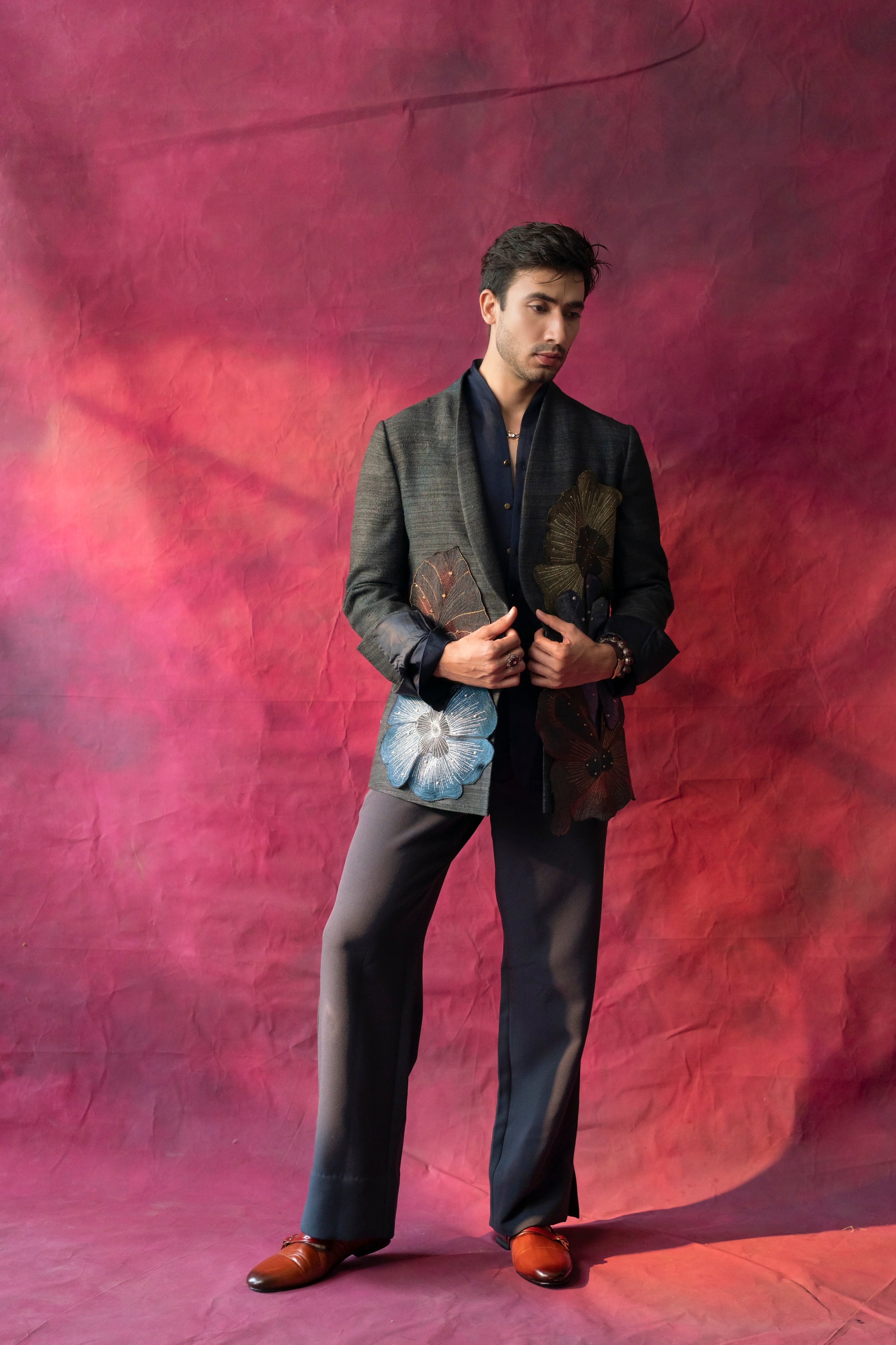 A stylish ensemble featuring a raw silk blazer in charcoal grey, adorned with multi-color hand-embroidered flowers placed in an abstract design. Paired with a linen silk shirt and matching trousers for a refined look.