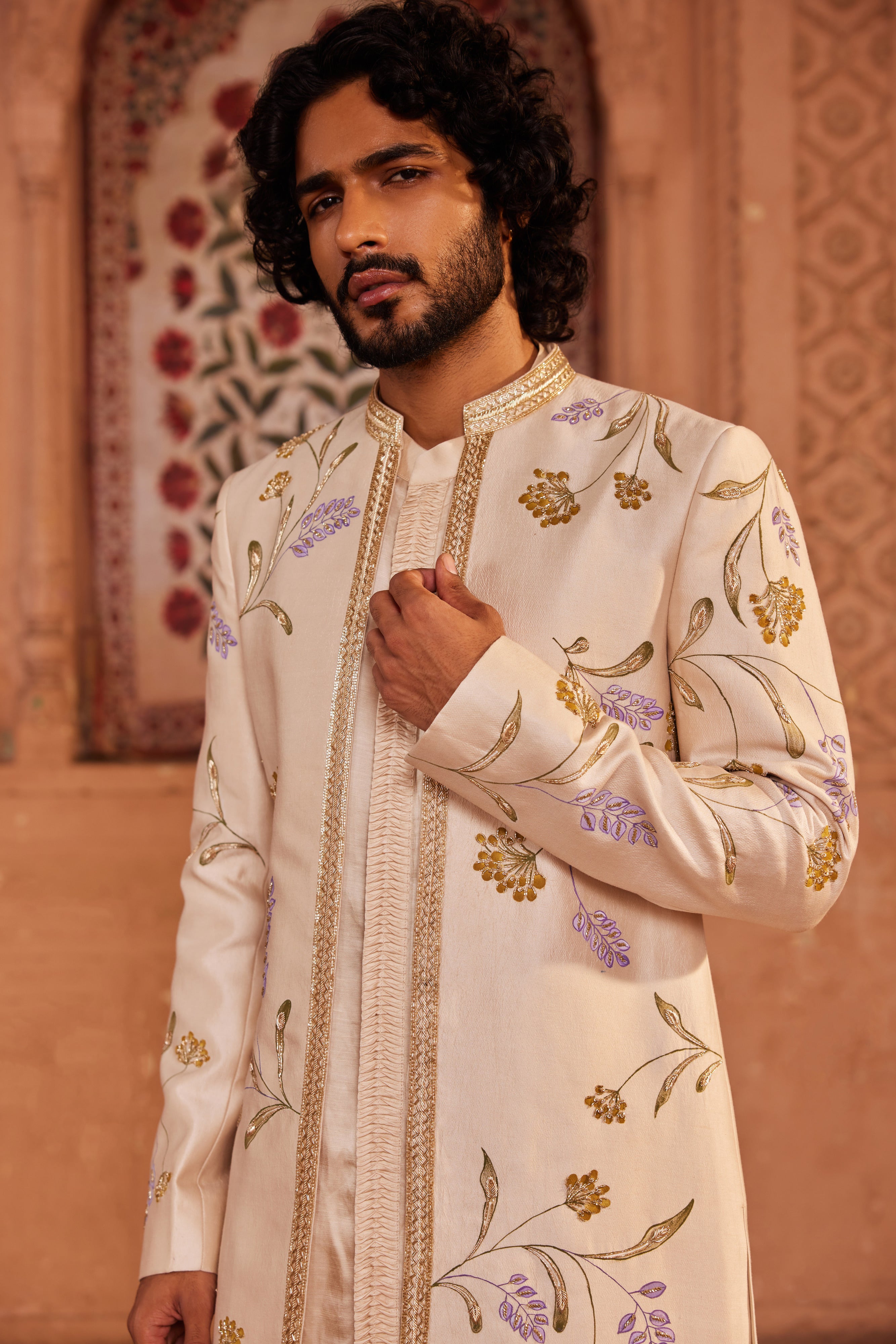 An open sherwani set featuring delicate hand-painted floral designs, embellished with dabka, zardozi, and cutdana work, paired with a kurta showcasing detailed fabric manipulation on the placket and matching pants.