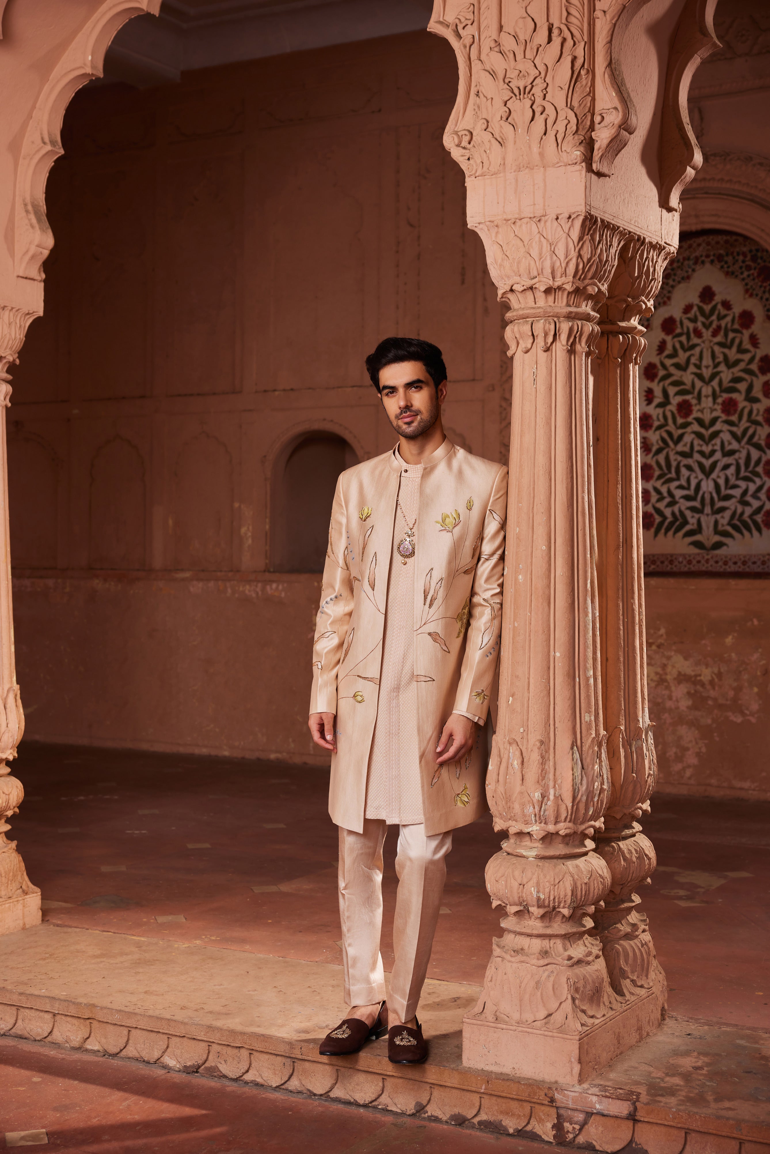 The Bageecha Open Sherwani Set showcases a sherwani adorned with meticulously hand-painted designs and detailed dabka and zardozi embellishments. It is complemented by a simple, monotone kurta and pants