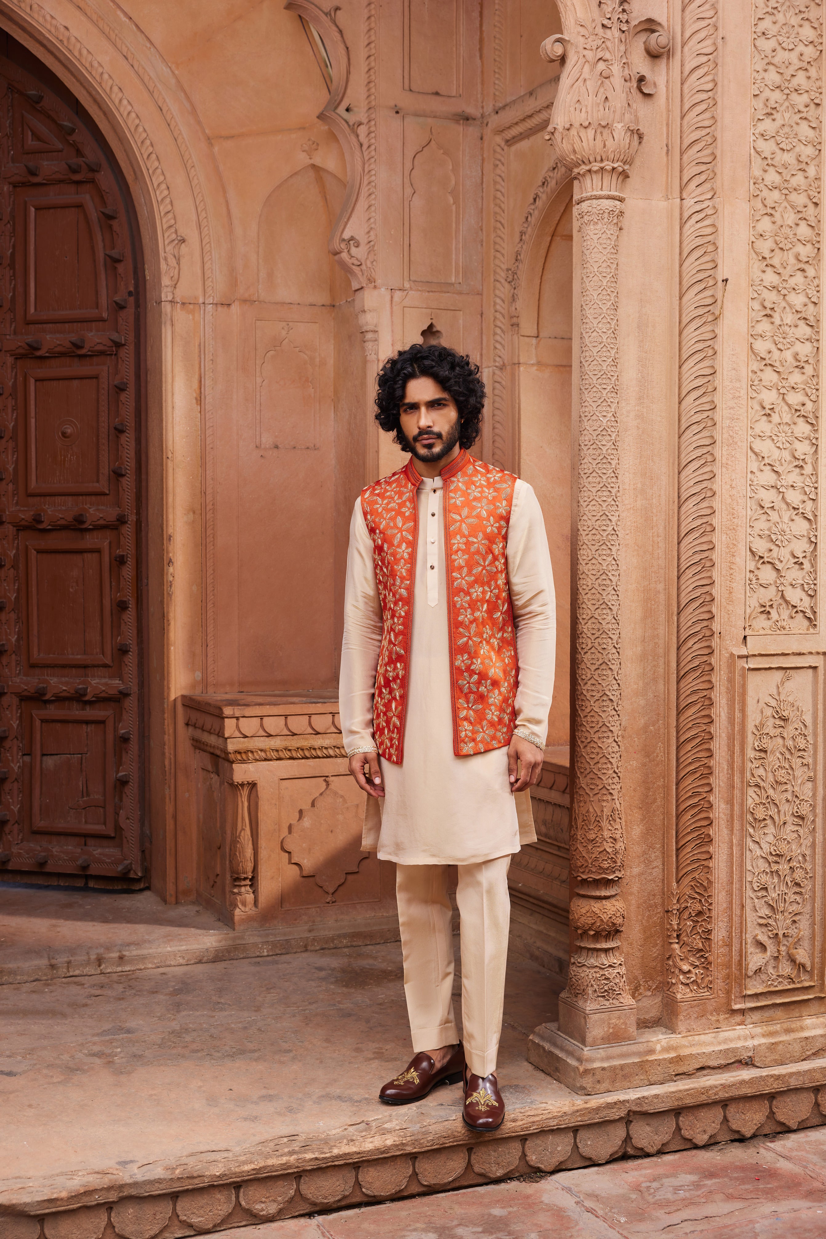 A Rust Floral Nehru Jacket Set featuring a rust-colored jacket adorned with clusters of gold floral motifs and dori embellishments. Paired with a contrast beige kurta detailed at the hem and matching pants.