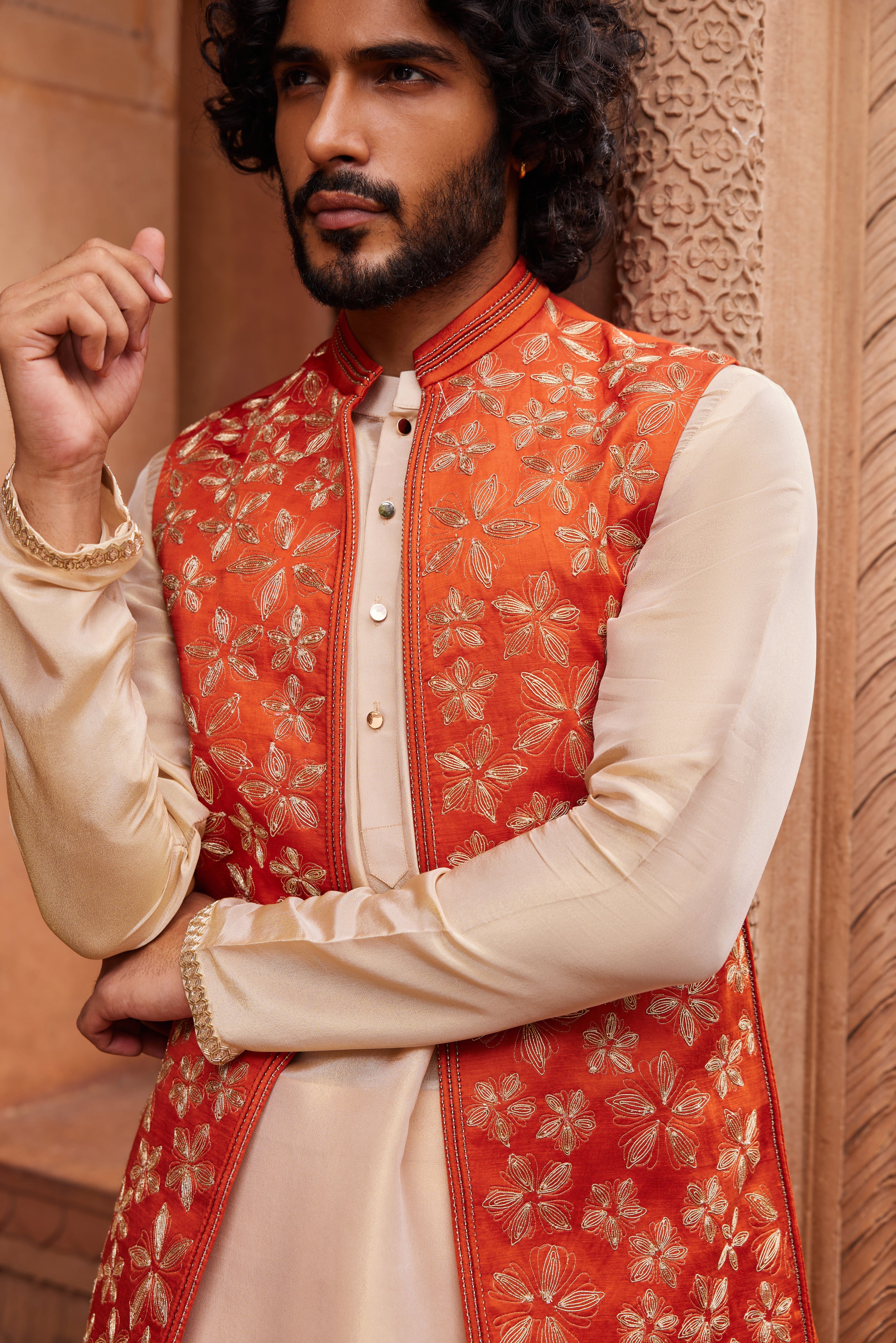 The Rust Floral Nehru Jacket Set showcases a rust jacket embellished with intricate gold floral clusters and dori work, complemented by a beige kurta with detailed hem and paired with pants